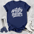 Navy blue t-shirt with 'artistry matters' text, silver tumbler, gloves, and white skates on a textured gray background