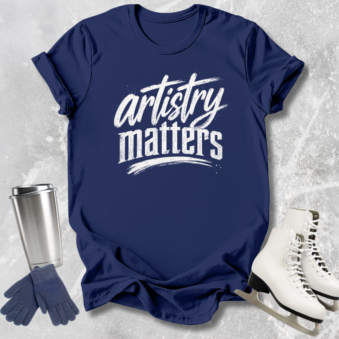 Navy blue t-shirt with 'artistry matters' text, silver tumbler, gloves, and white skates on a textured gray background