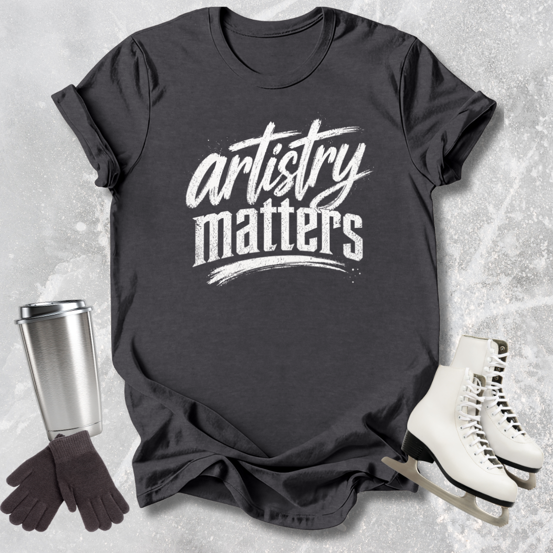 Black t-shirt with 'artistry matters' text, silver tumbler, black gloves, and white ice skates on a textured gray background
