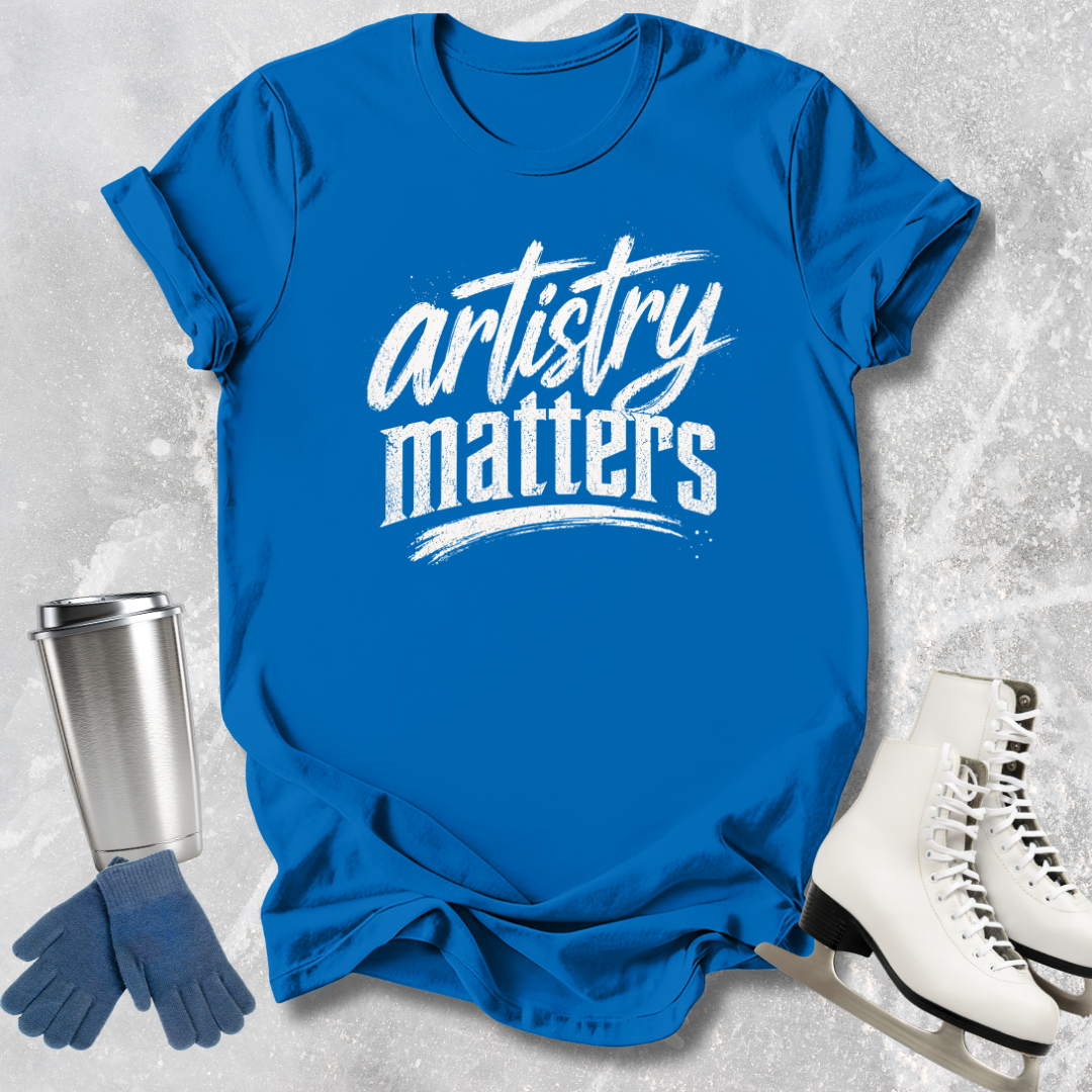 Blue t-shirt with 'artistry matters' text, silver tumbler, blue gloves, and white ice skates on a textured gray background