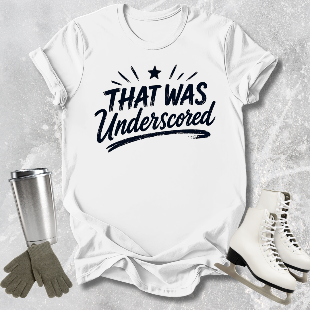 White t-shirt with 'THAT WAS Underscored' text, next to a silver tumbler, gray gloves, and white skates on a light gray background.
