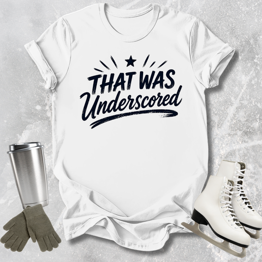 White t-shirt with 'THAT WAS Underscored' text, next to a silver tumbler, gray gloves, and white skates on a light gray background.