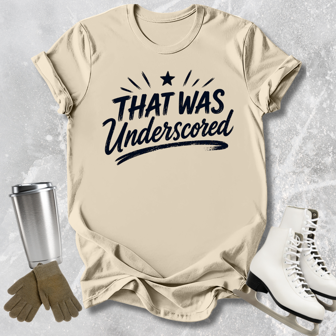 Beige t-shirt with 'THAT WAS Underscored' text, next to a silver tumbler, brown gloves, and white skates on a textured gray background.