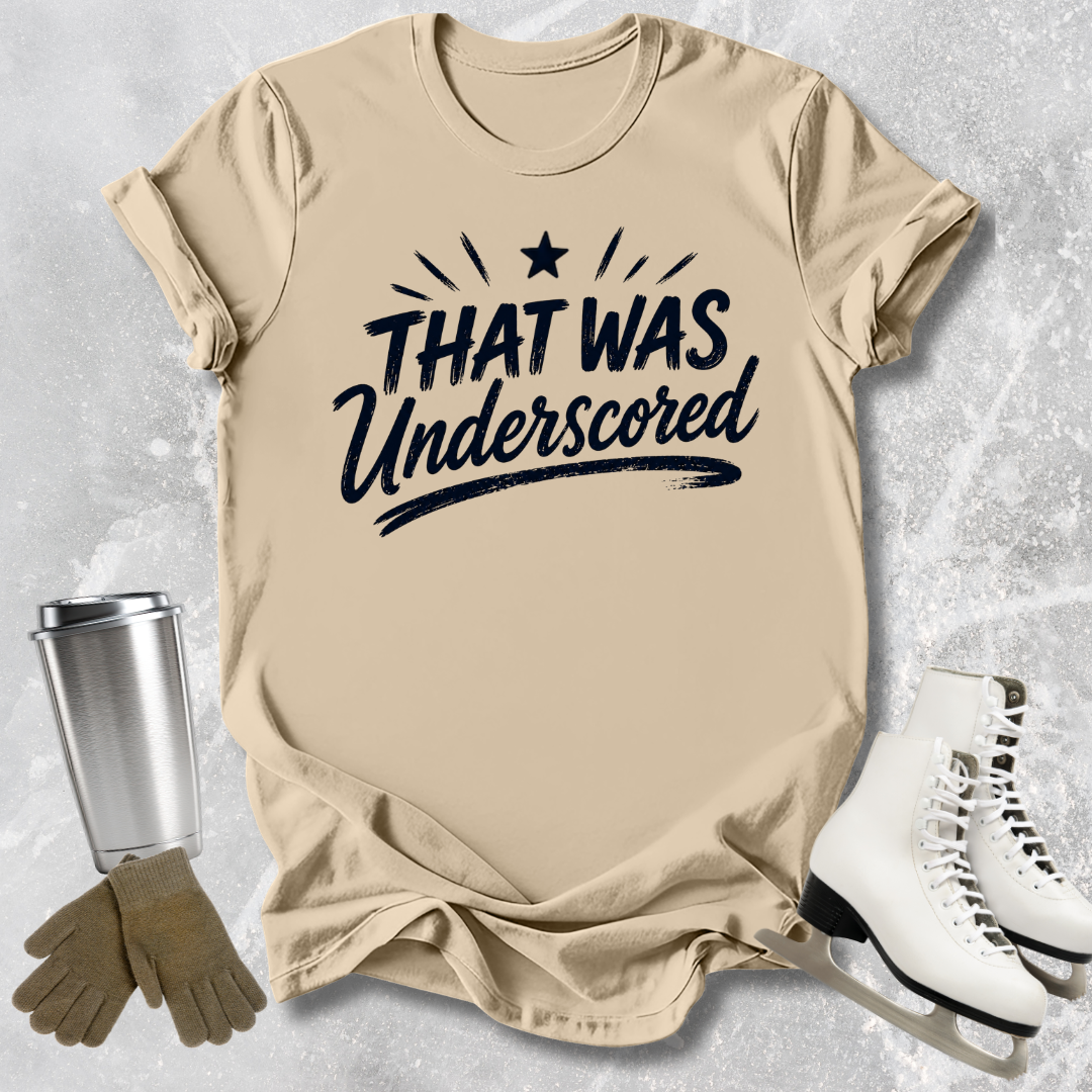 Beige t-shirt with 'THAT WAS Underscored' text, next to a silver tumbler, brown gloves, and white skates on a textured gray background.