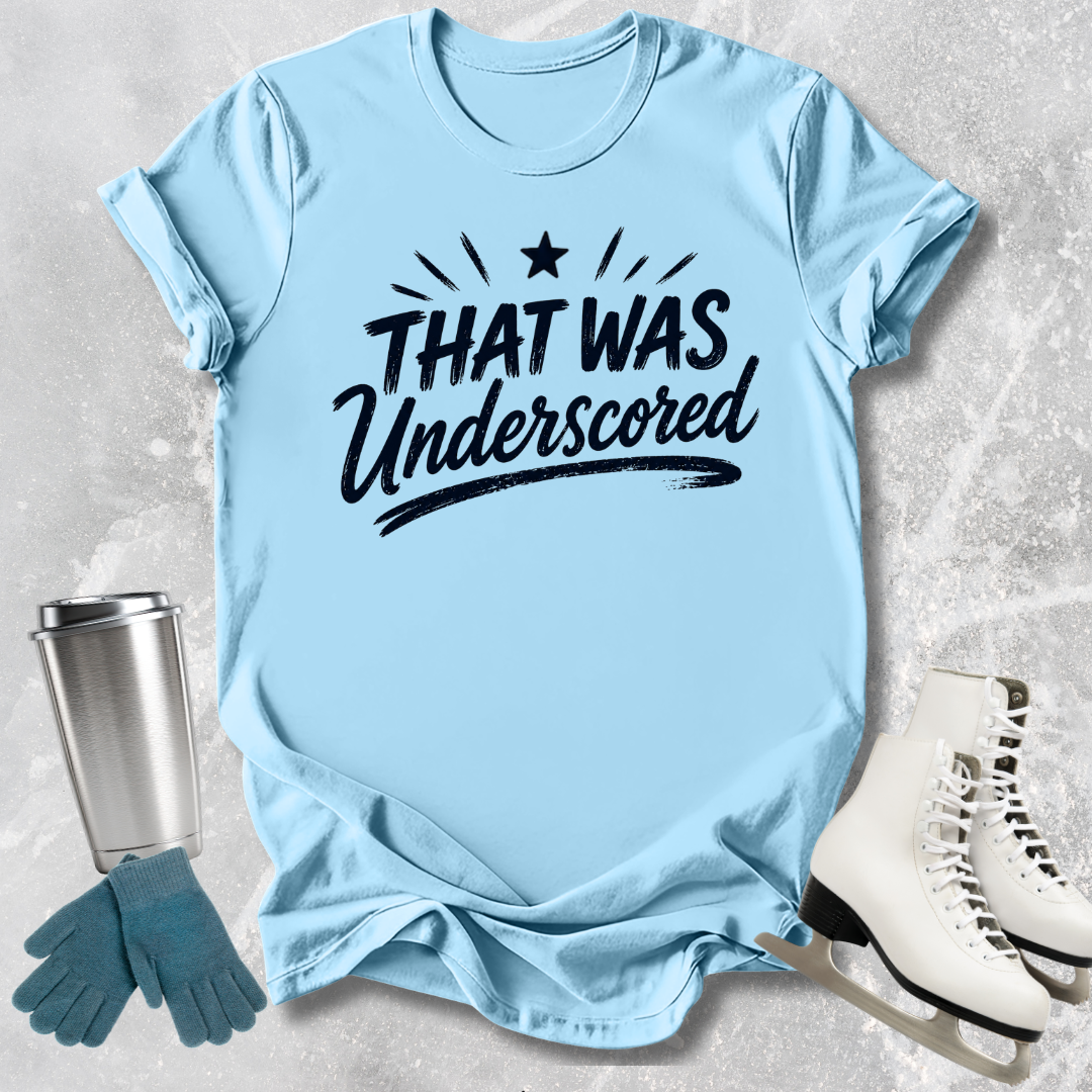 Light blue t-shirt with 'THAT WAS Underscored' text, next to a silver tumbler, green gloves, and white skates on a textured surface.