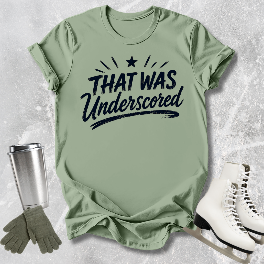 Green t-shirt with 'THAT WAS Underscored' text, next to a silver tumbler and white skates on a textured surface.