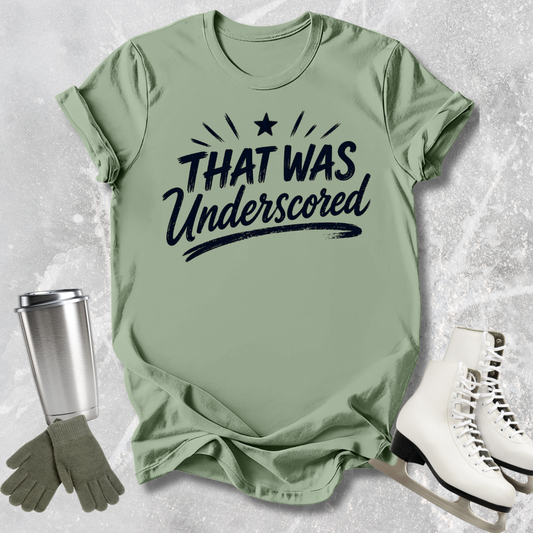 Green t-shirt with 'THAT WAS Underscored' text, next to a silver tumbler and white skates on a textured surface.