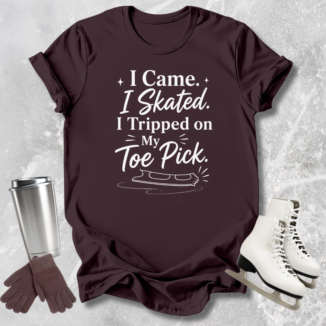 Maroon t-shirt with humorous ice skating quote 'I came I skated. I tripped on my toe pick'  next to ice skates and a tumbler on a textured surface.