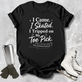 Black t-shirt with 'I came I skated. I tripped on my toe pick' text and ice skate graphic on a textured surface with ice skates and gloves.