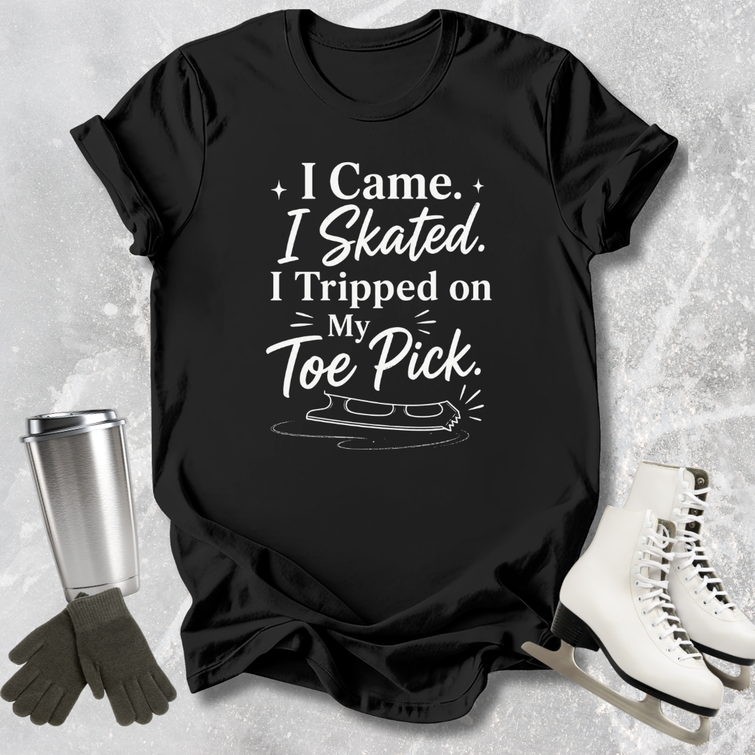 Black t-shirt with 'I came I skated. I tripped on my toe pick' text and ice skate graphic on a textured surface with ice skates and gloves.