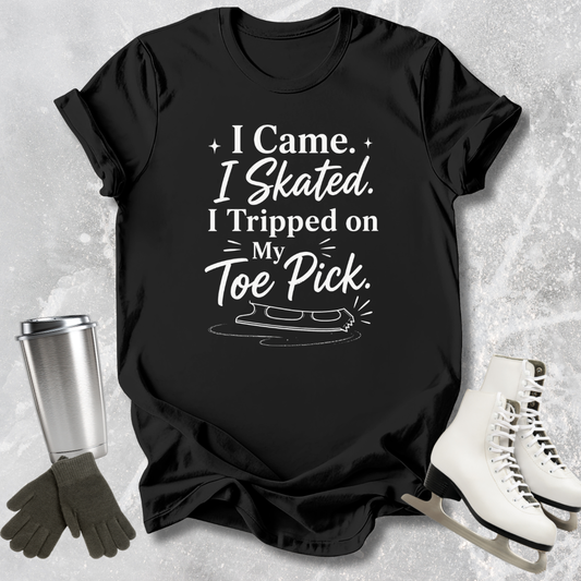 Black t-shirt with 'I came I skated. I tripped on my toe pick' text and ice skate graphic on a textured surface with ice skates and gloves.