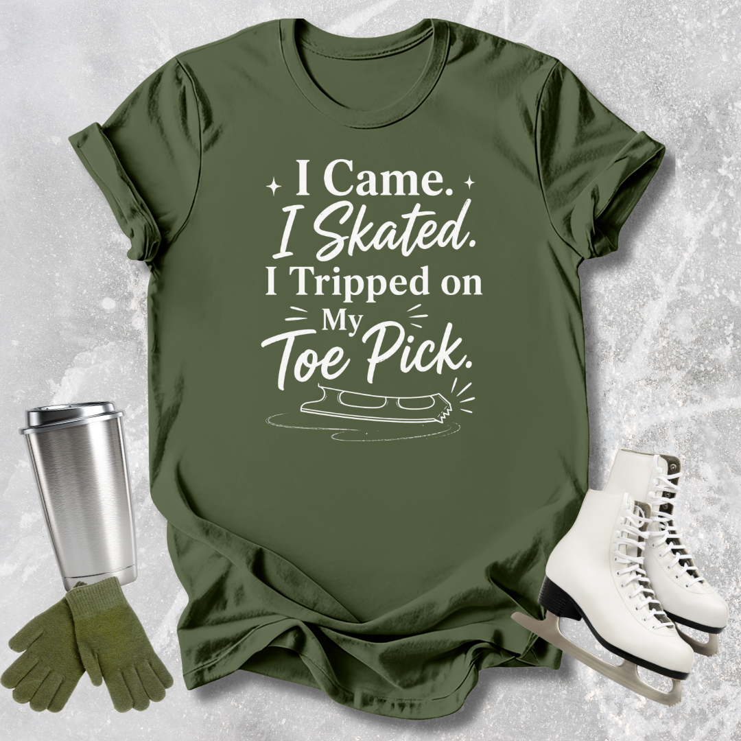 Green t-shirt with humorous 'I came I skated. I tripped on my toe pick' text, next to a silver tumbler, green gloves, and white ice skates on a textured gray background.