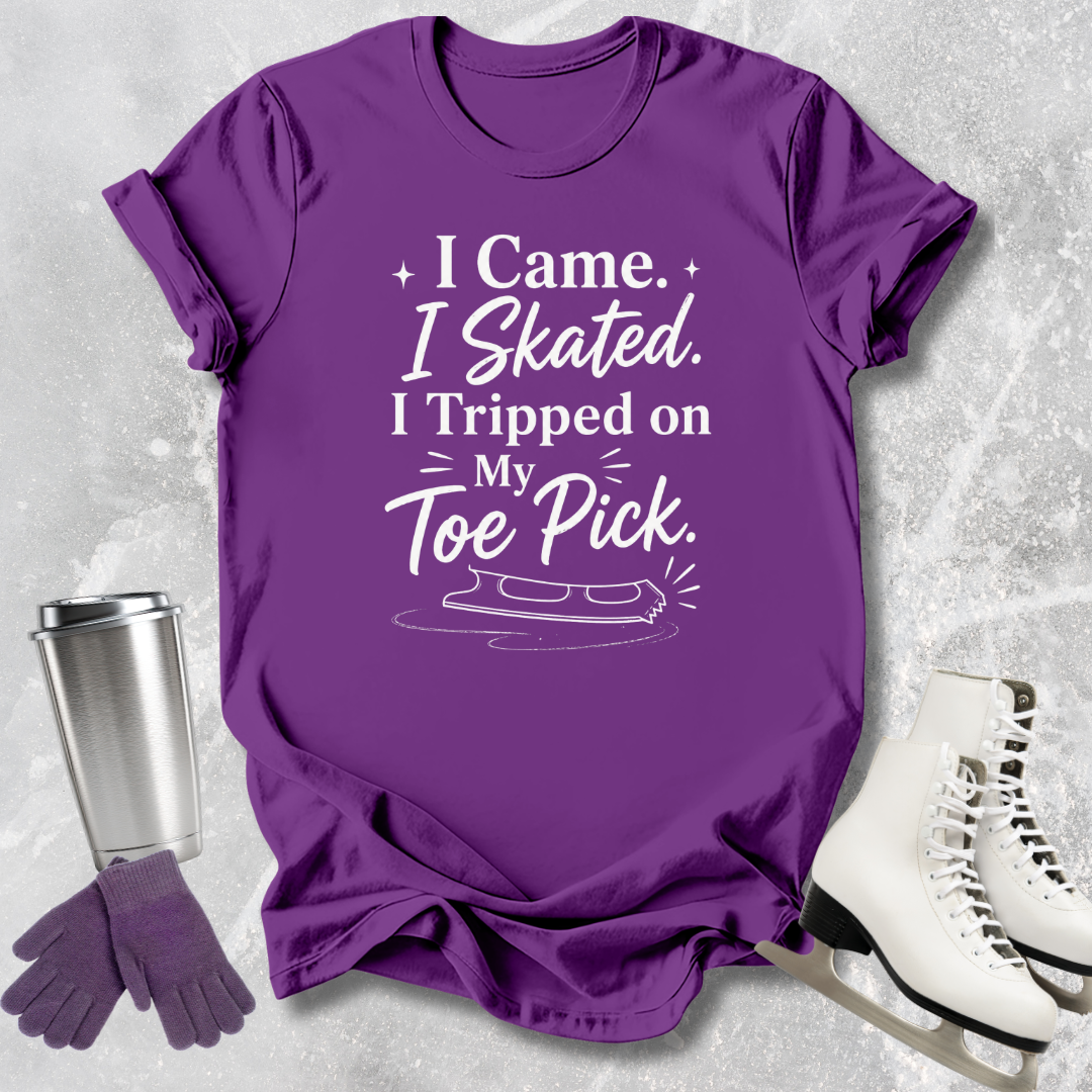 Purple t-shirt with humorous ice skating 'I came I skated. I tripped on my toe pick' text, next to ice skates and a tumbler on a textured surface.