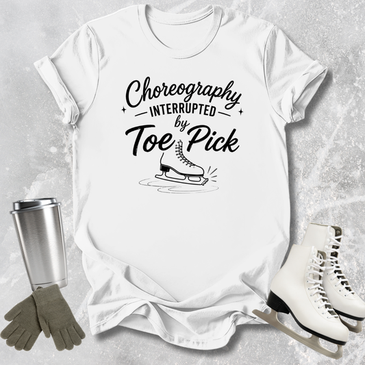 White t-shirt with 'Choreography Interrupted by Toe Pick' text, ice skates, gloves, and a tumbler on a gray background