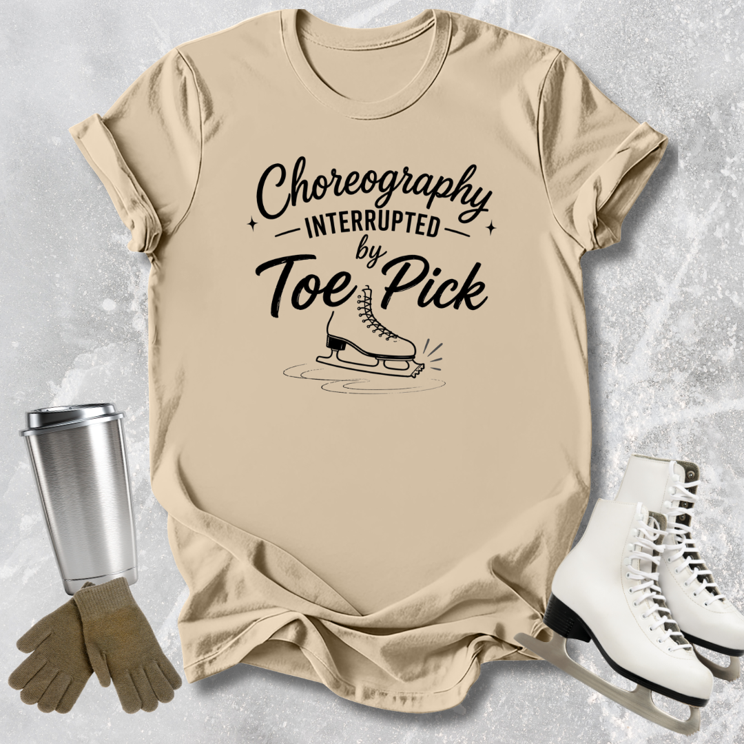 Beige t-shirt with 'Choreography Interrupted by Toe Pick' text and graphic, next to ice skates and a tumbler.