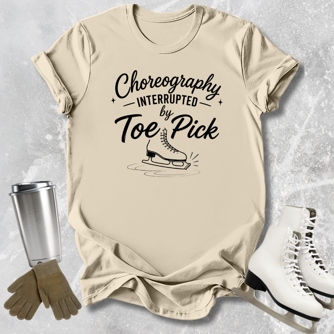 Beige t-shirt with 'Choreography Interrupted by Toe Pick' text and graphic, next to ice skates and a tumbler.
