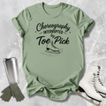 Green t-shirt with 'Choreography Interrupted by Toe Pick' text, ice skates, gloves, and a tumbler on a textured surface.