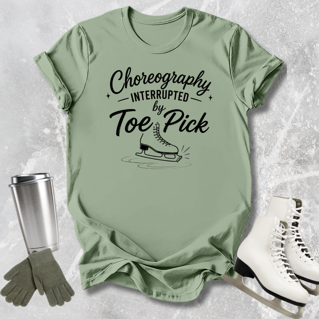 Green t-shirt with 'Choreography Interrupted by Toe Pick' text, ice skates, gloves, and a tumbler on a textured surface.