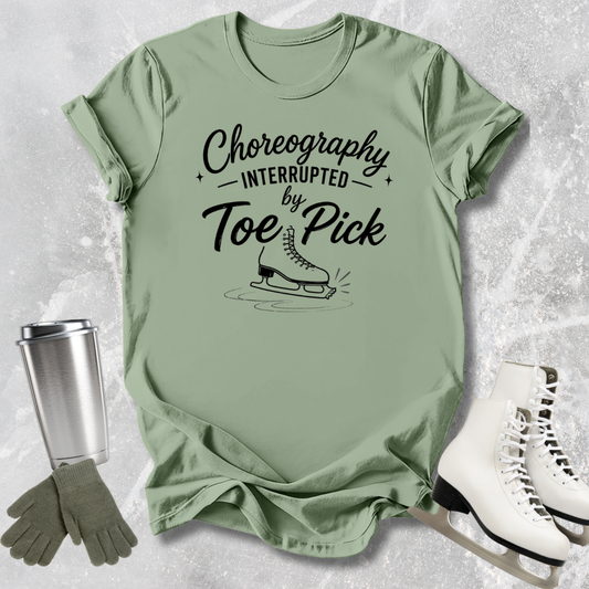 Green t-shirt with 'Choreography Interrupted by Toe Pick' text, ice skates, gloves, and a tumbler on a textured surface.