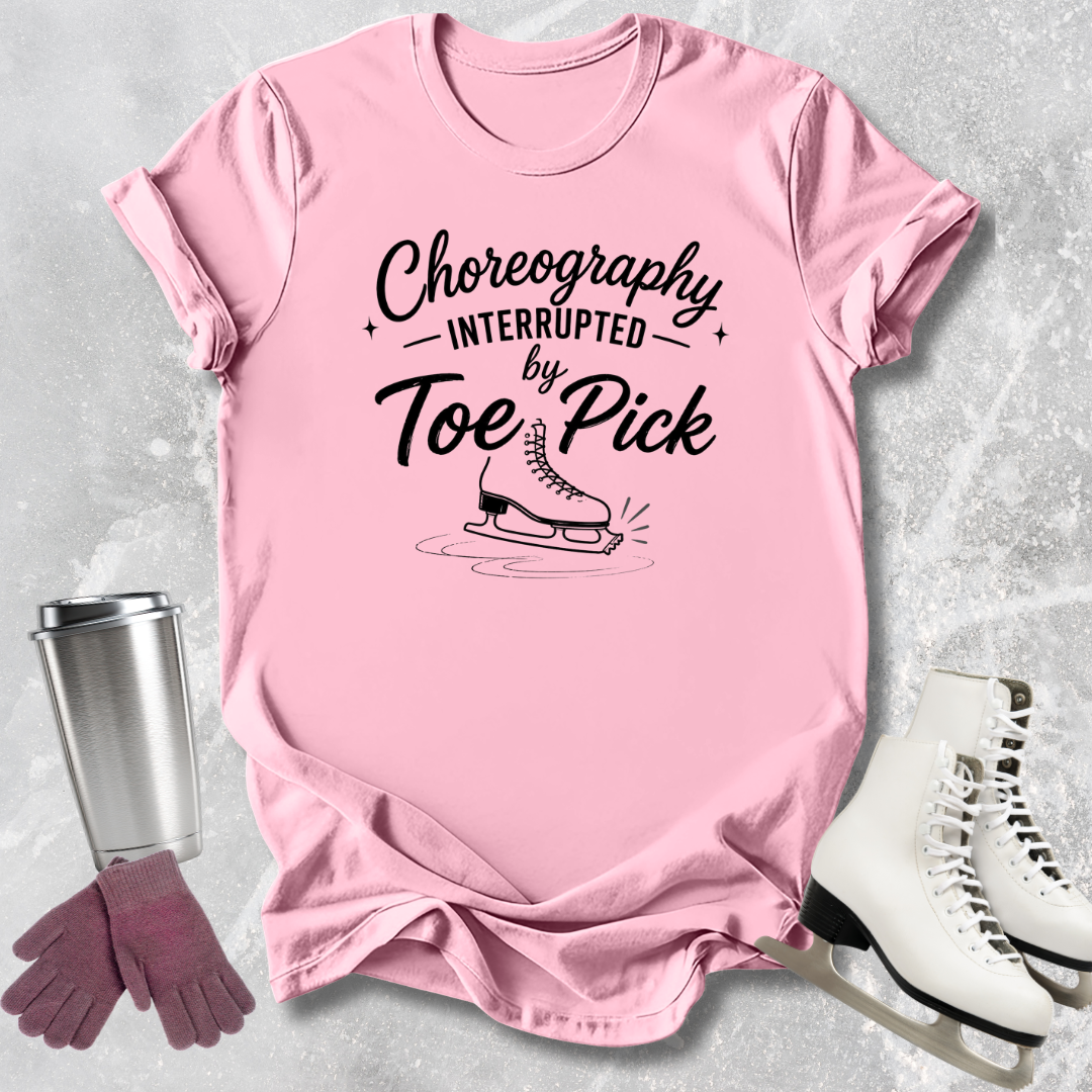 Pink t-shirt with 'Choreography Interrupted by Toe Pick' text, next to ice skates and a tumbler on a textured surface.