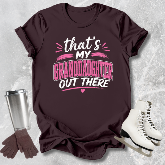 Maroon t-shirt with 'That's my granddaughter out there' text, next to a silver tumbler, brown gloves, and white skates on a textured gray background.