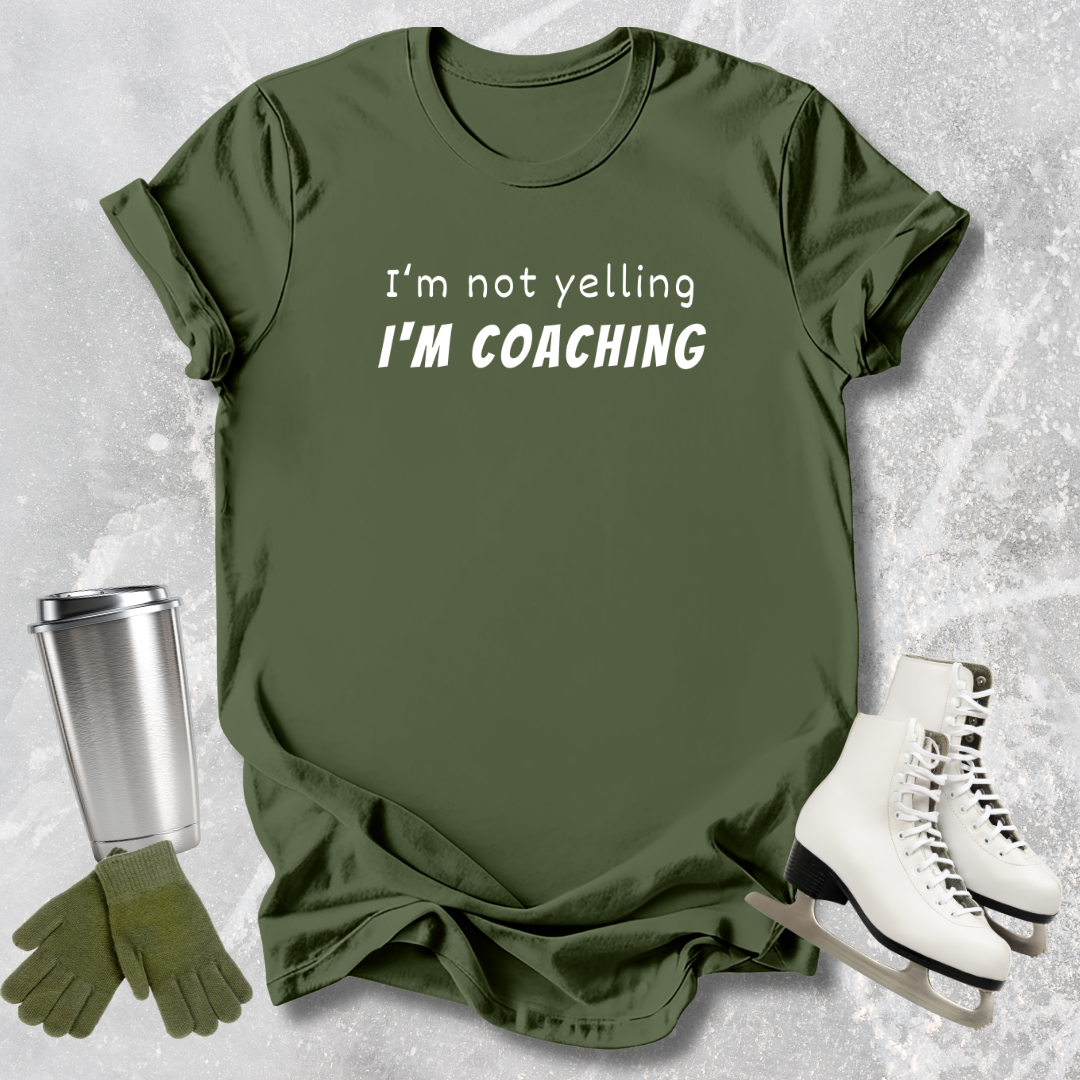Green t-shirt with 'I'm not yelling I'm coaching' text, next to a silver tumbler, green gloves, and white skates on a textured gray background.