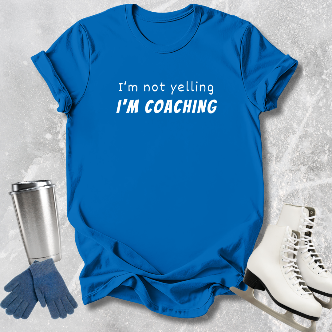 Blue t-shirt with 'I'm not yelling I'm coaching' text, next to a silver tumbler, blue gloves, and white skates on a textured gray background.