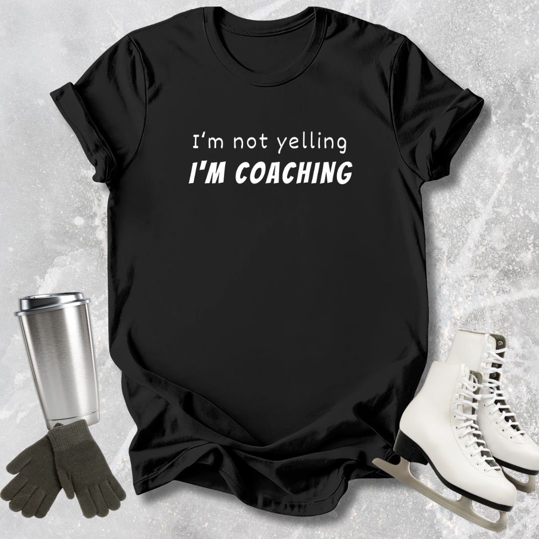 Black t-shirt with 'I'm not yelling I'm coaching' text, surrounded by ice skates and a tumbler on a textured surface.