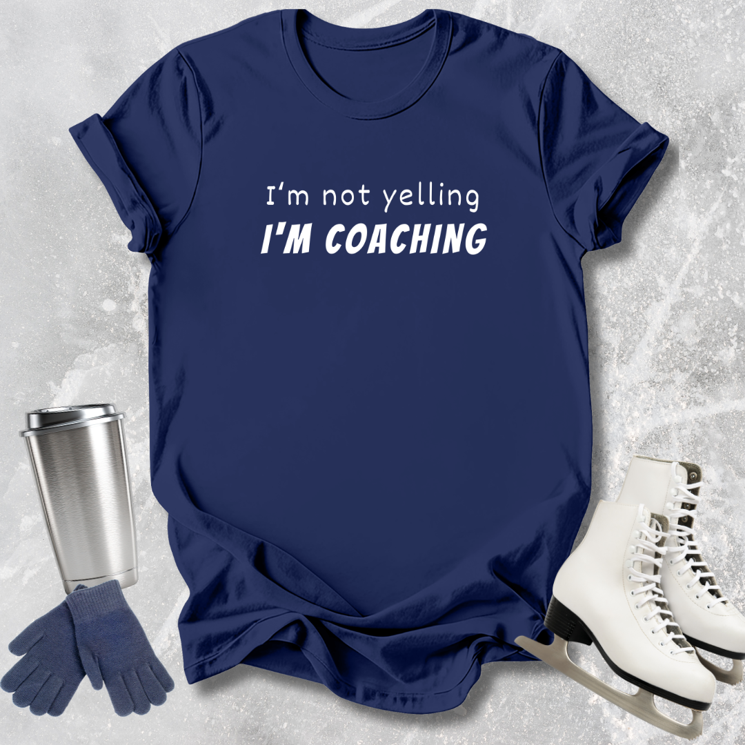 Navy blue t-shirt with 'I'm not yelling I'm coaching' text, next to a silver tumbler, gloves, and white skates on a light gray textured background.