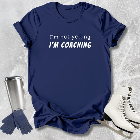 Navy blue t-shirt with 'I'm not yelling I'm coaching' text, next to a silver tumbler, gloves, and white skates on a light gray textured background.