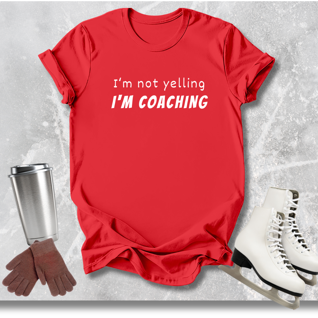 Red t-shirt with 'I'm not yelling I'm coaching' text, next to a silver tumbler, brown gloves, and white skates on a light gray background.