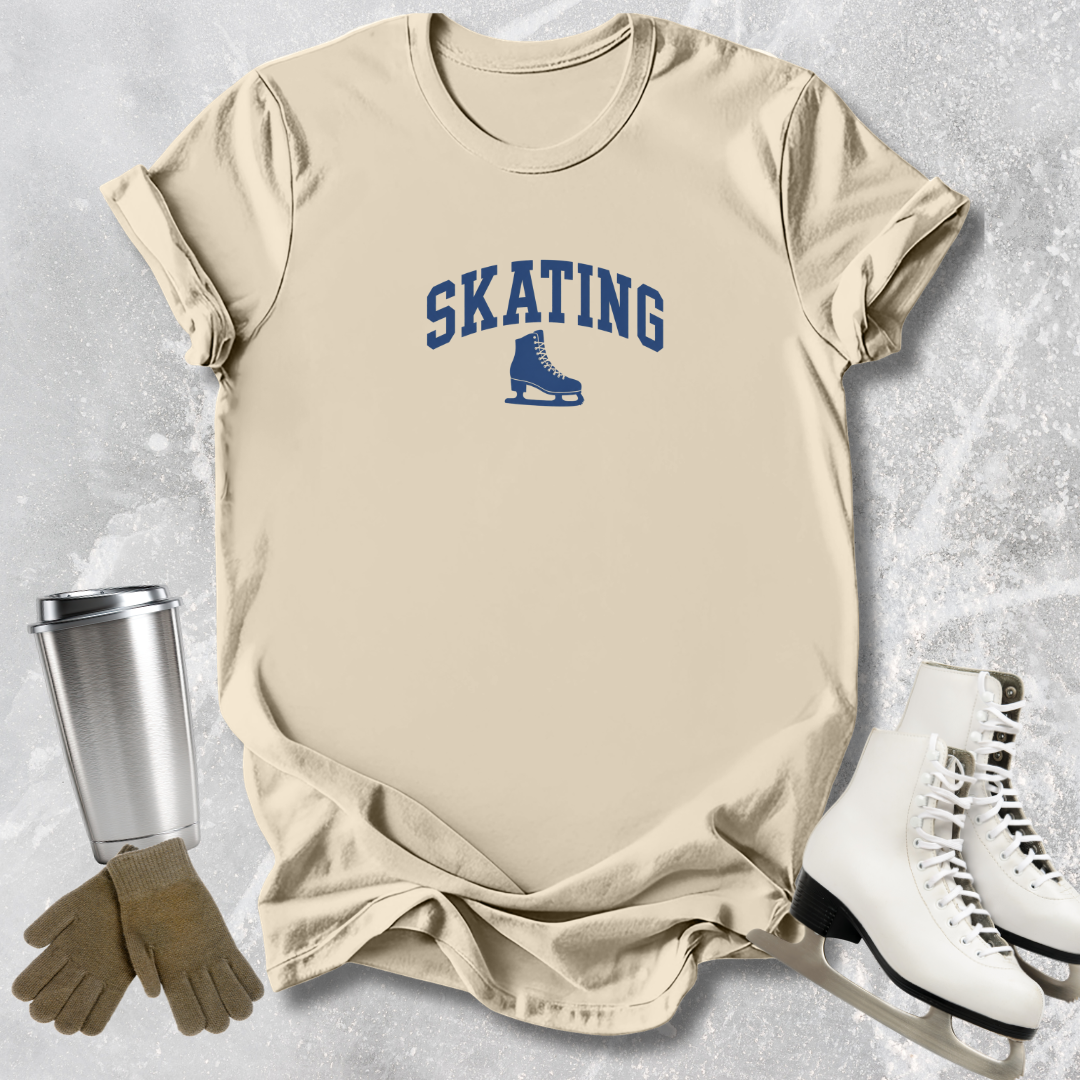 Beige t-shirt with 'SKATING' text and ice skate graphic, next to a silver tumbler, brown gloves, and white skates on a textured gray background.