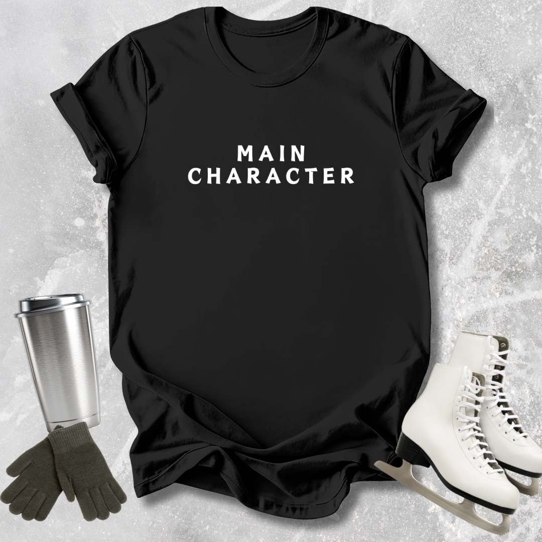 Black t-shirt with 'MAIN CHARACTER' text, next to ice skates and a tumbler on a textured surface.