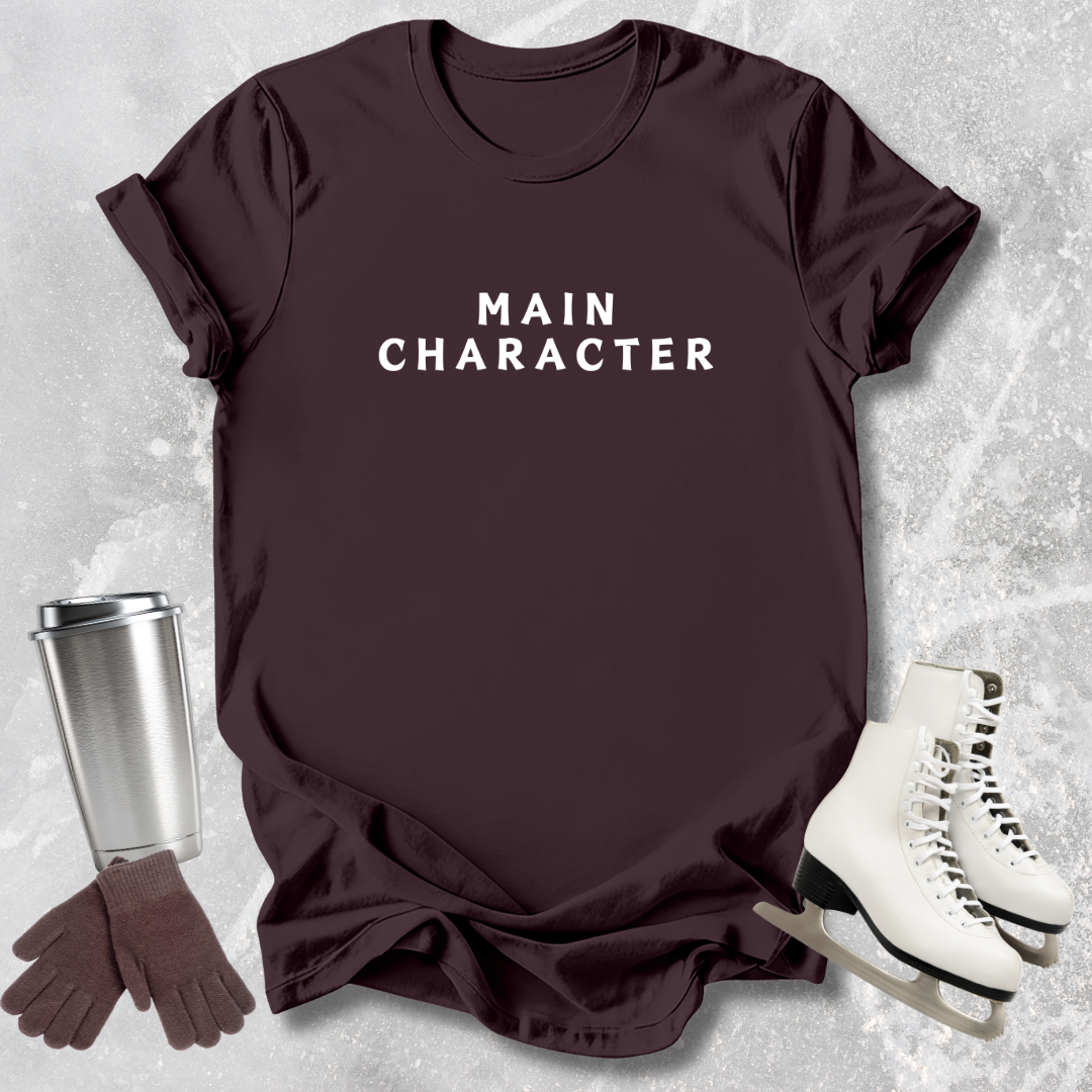 Maroon t-shirt with 'MAIN CHARACTER' text, silver tumbler, brown gloves, and white skates on a textured surface.