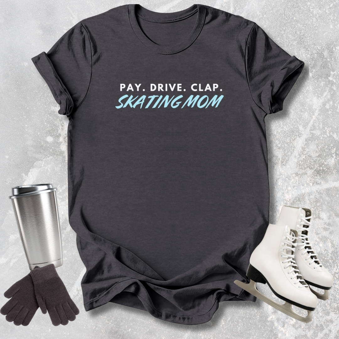 Dark gray t-shirt with 'Maroon t-shirt with 'PAY. DRIVE. Clap Figure Skating Mom ' text, next to a silver tumbler, black gloves, and white skates on a textured surface.