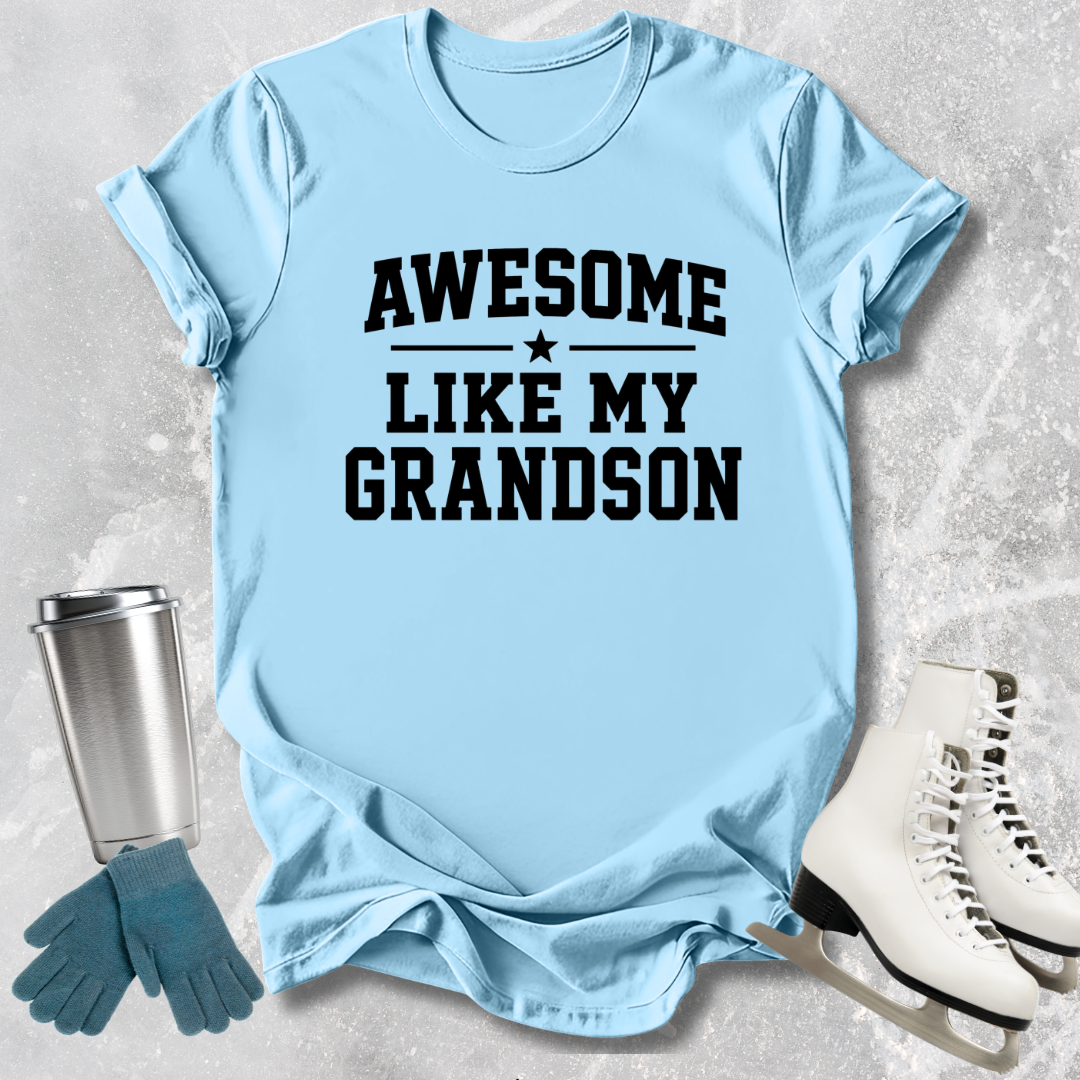 Light blue t-shirt with 'Awesome like my grandson' text, next to a silver tumbler, green gloves, and white skates on a textured surface.