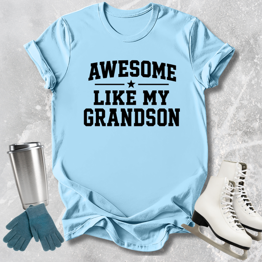 Light blue t-shirt with 'Awesome like my grandson' text, next to a silver tumbler, green gloves, and white skates on a textured surface.