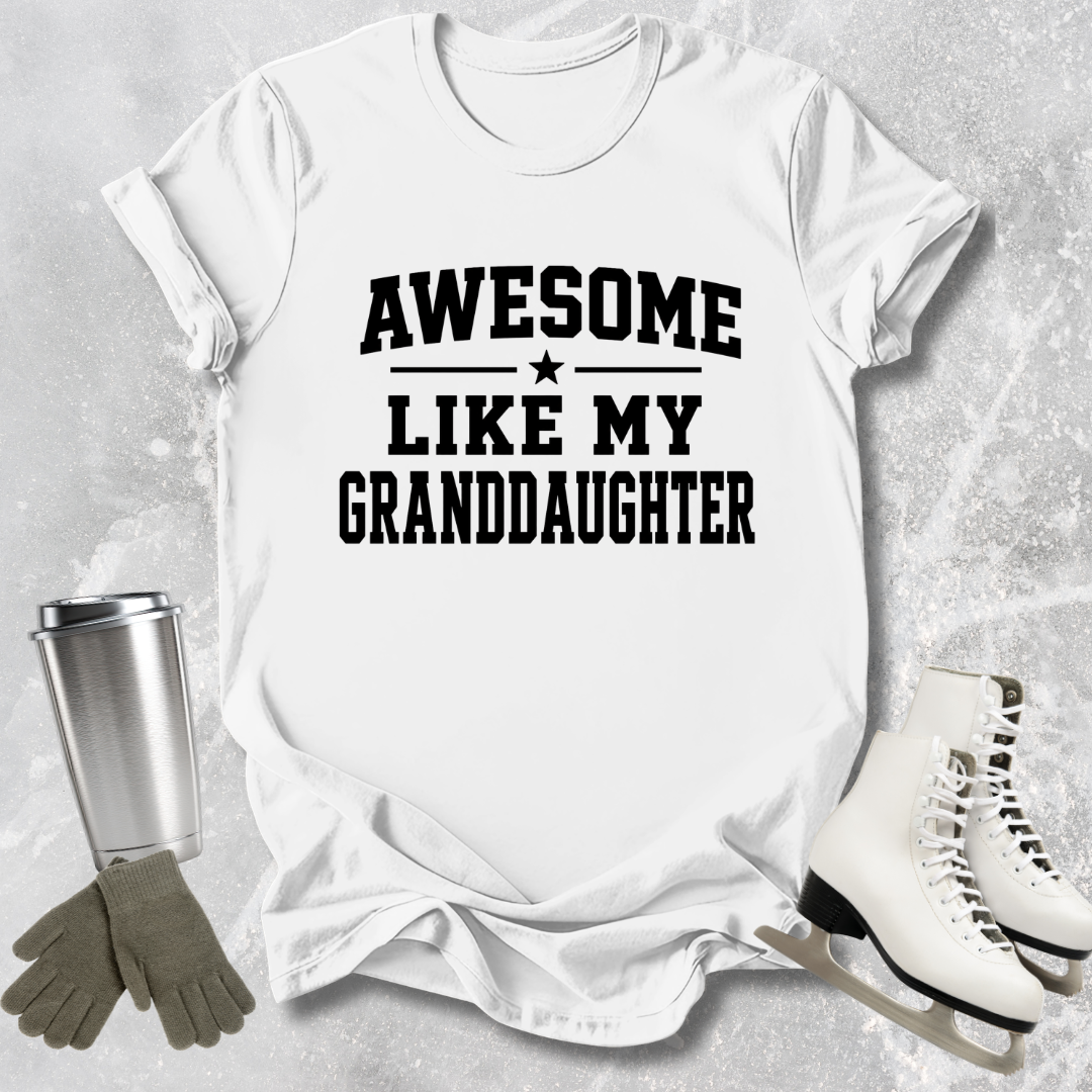 White t-shirt with 'Awesome like my granddaughter' text, next to a silver tumbler, gloves, and ice skates on a light gray background.