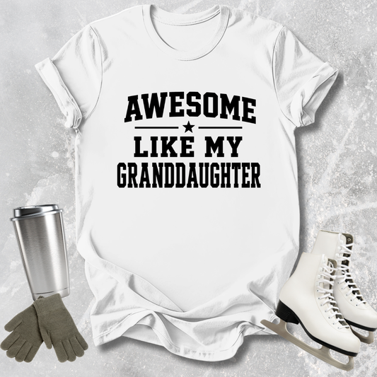 White t-shirt with 'Awesome like my granddaughter' text, next to a silver tumbler, gloves, and ice skates on a light gray background.
