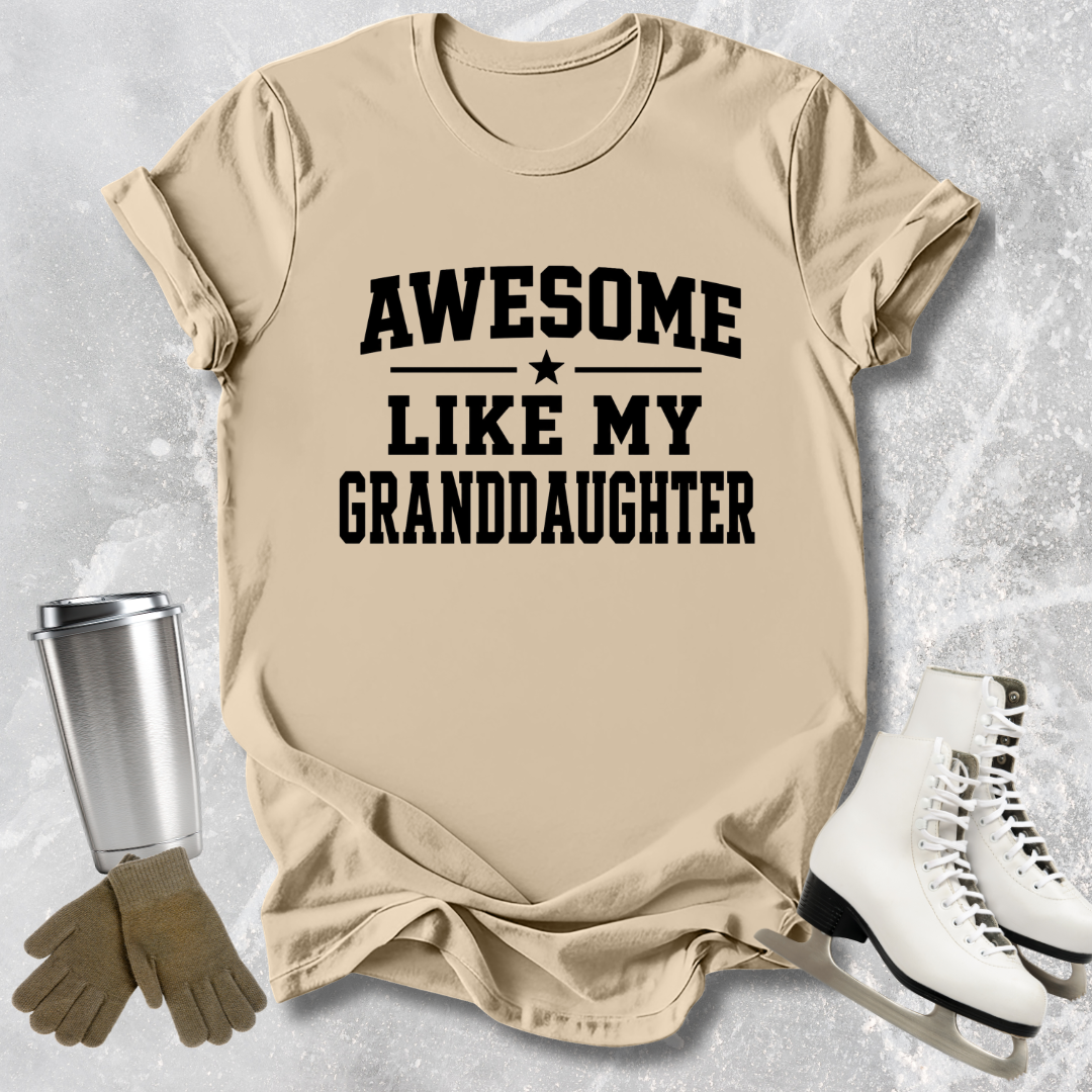 Beige t-shirt with 'Awesome like my granddaughter' text, next to a silver tumbler, brown gloves, and white ice skates on a light gray textured background.