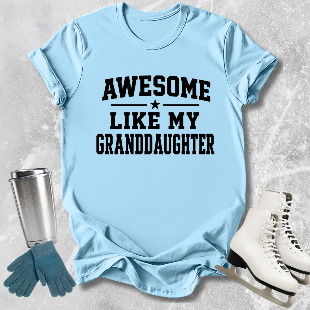 Light blue t-shirt with 'Awesome like my granddaughter' text, next to a silver tumbler, green gloves, and white skates on a textured surface.
