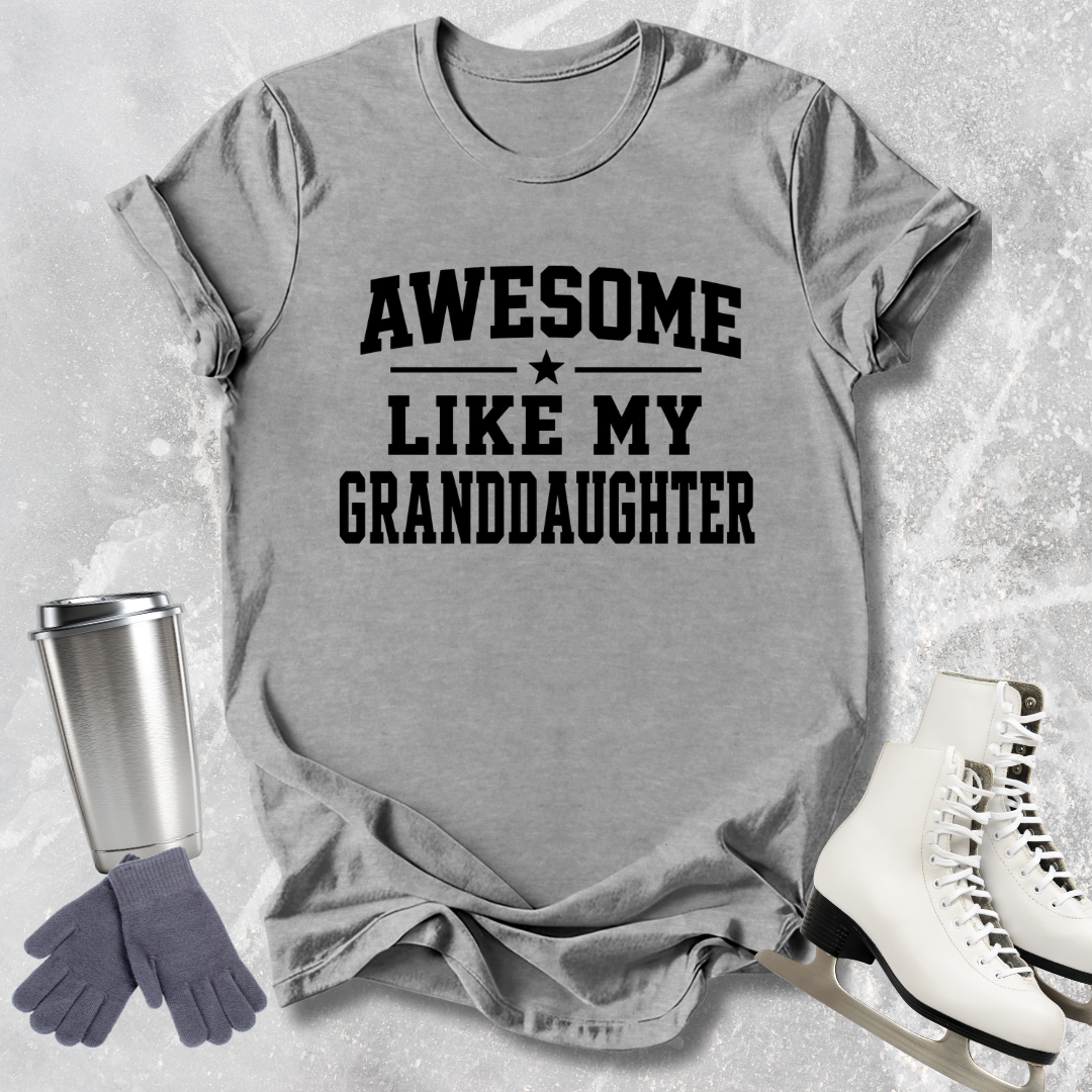 Gray t-shirt with 'Awesome like my granddaughter' text, next to a silver tumbler, gray gloves, and white skates on a textured surface.