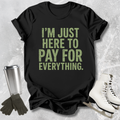 Black t-shirt with green text 'I'm just here to pay for everything' on a textured surface with accessories.