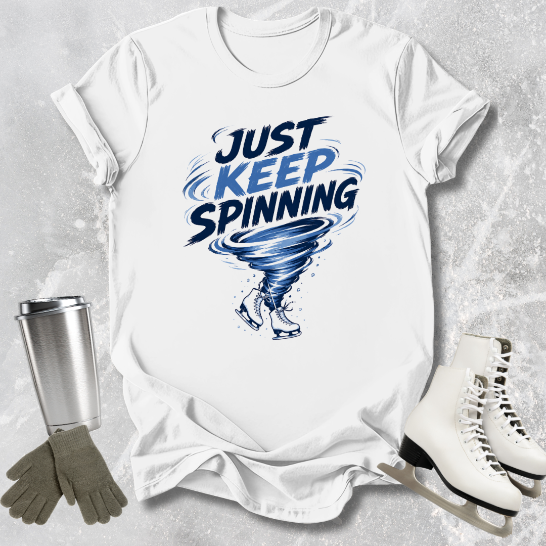 White t-shirt with 'Just Keep Spinning' graphic, silver tumbler, gloves, and ice skates on a textured gray background