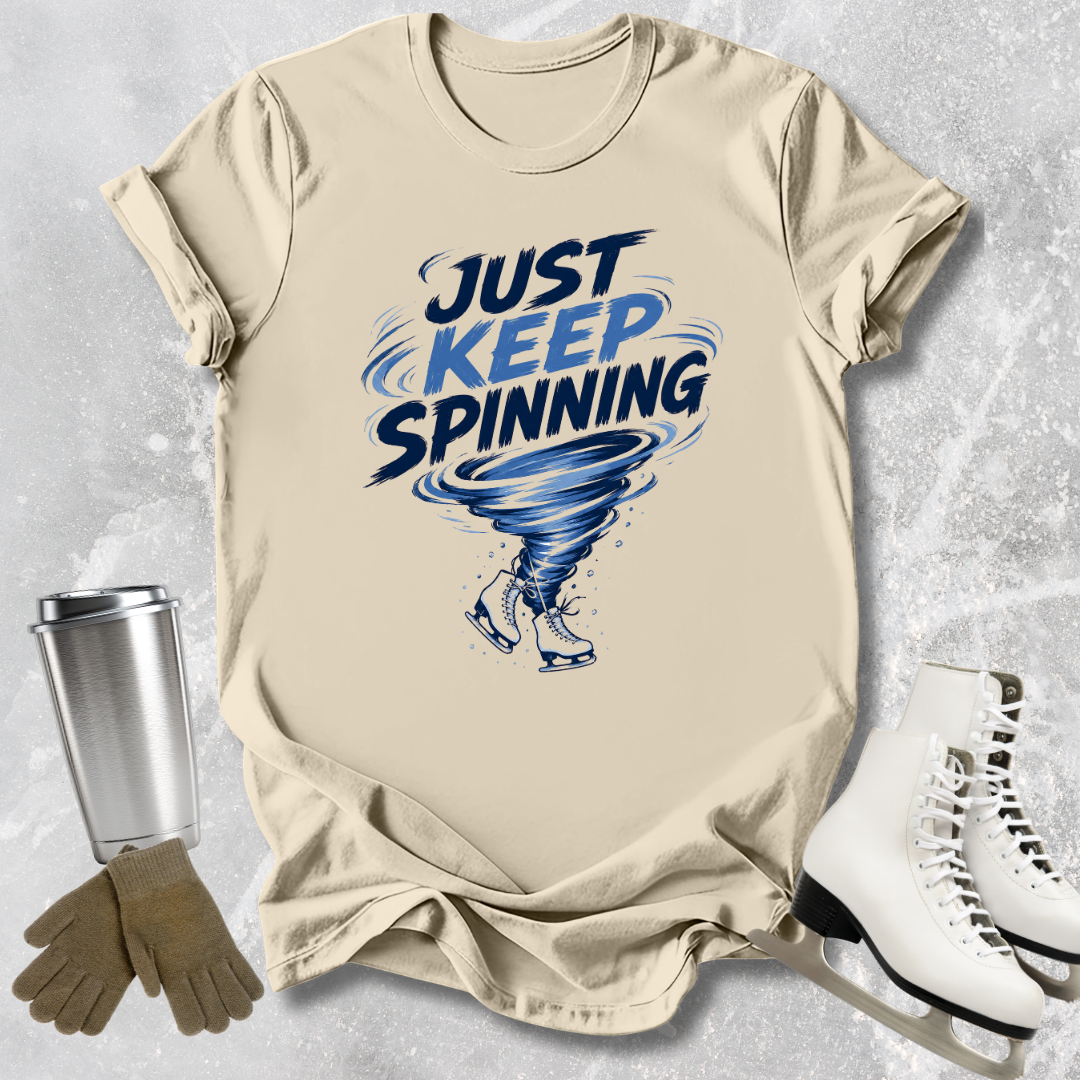 Beige t-shirt with 'Just Keep Spinning' graphic, silver tumbler, brown gloves, and white skates on a textured gray background