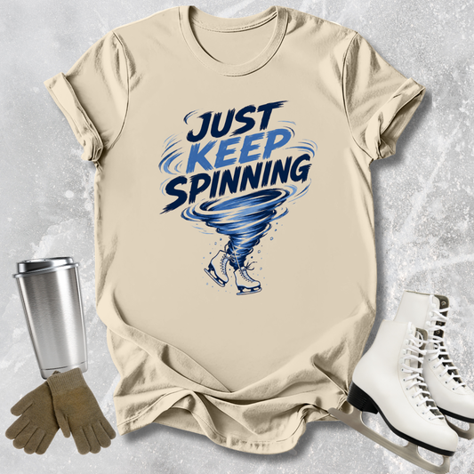Beige t-shirt with 'Just Keep Spinning' graphic, silver tumbler, brown gloves, and white skates on a textured gray background