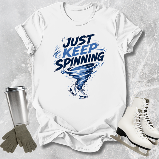 White t-shirt with 'Just Keep Spinning' graphic, silver tumbler, gloves, and ice skates on a textured gray background