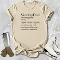 Beige t-shirt with 'Skating Dad' definition text, placed on a textured surface with ice skates and a tumbler.