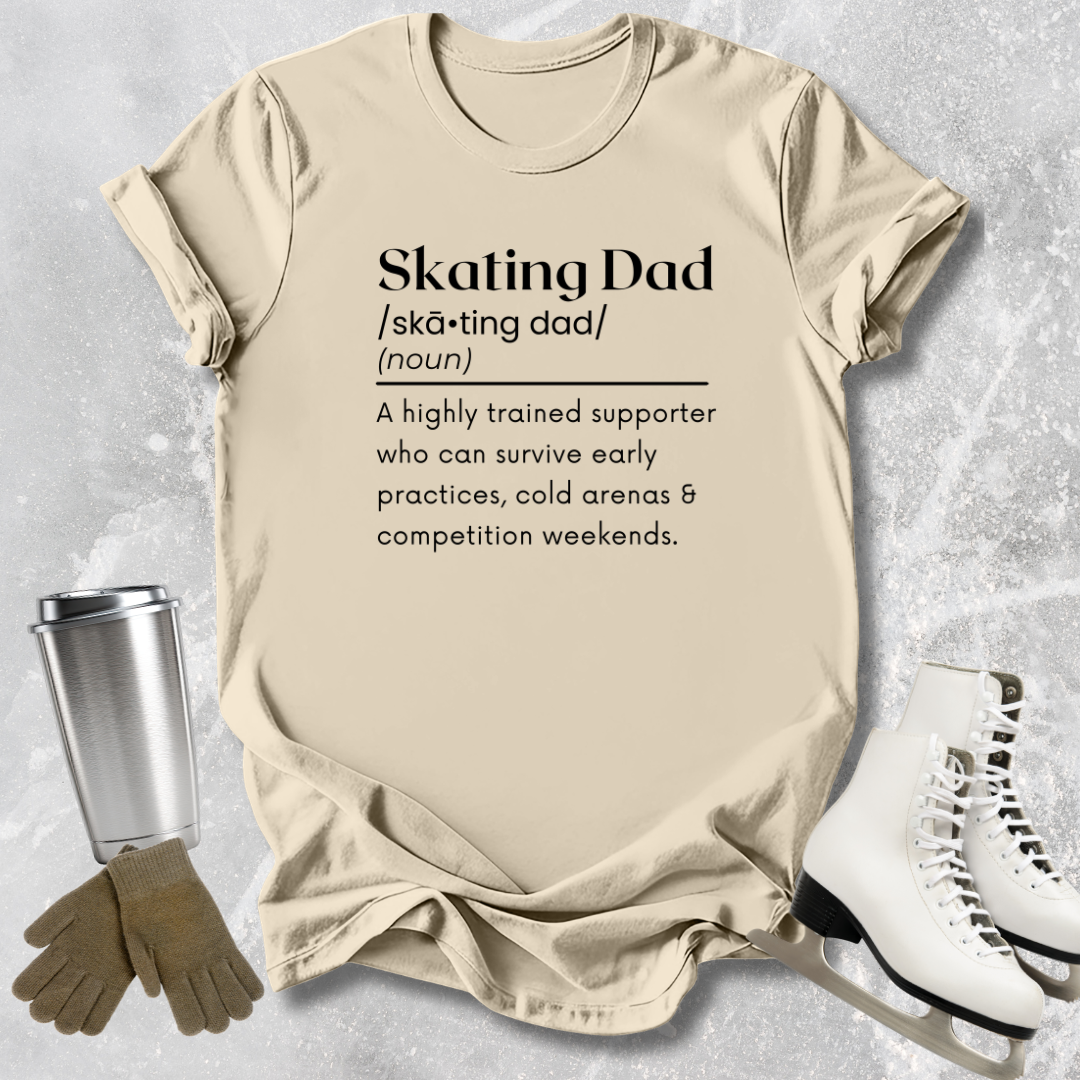 Beige t-shirt with 'Skating Dad' definition text, placed on a textured surface with ice skates and a tumbler.