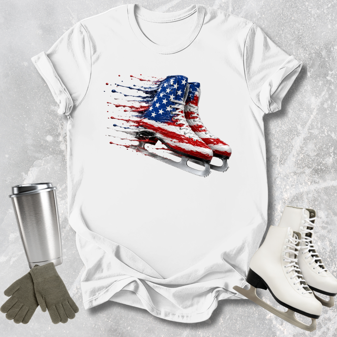 White t-shirt with American flag ice skate design, surrounded by ice skates and a tumbler on a textured surface.